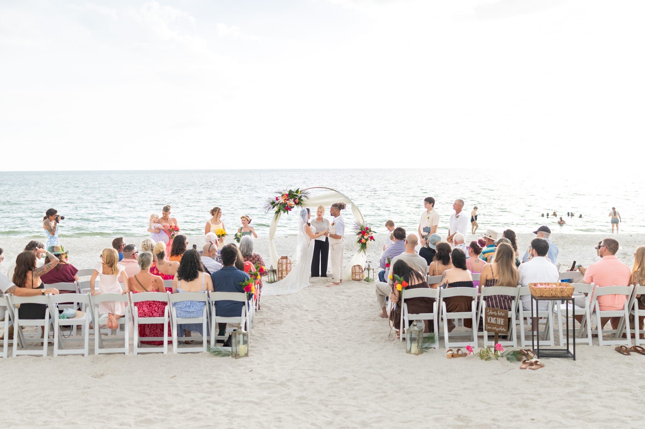 A full beach wedding with rows of seated guests watching a couple exchange vows with officiant Beth Schell under a circular arch at the edge of the Gulf.