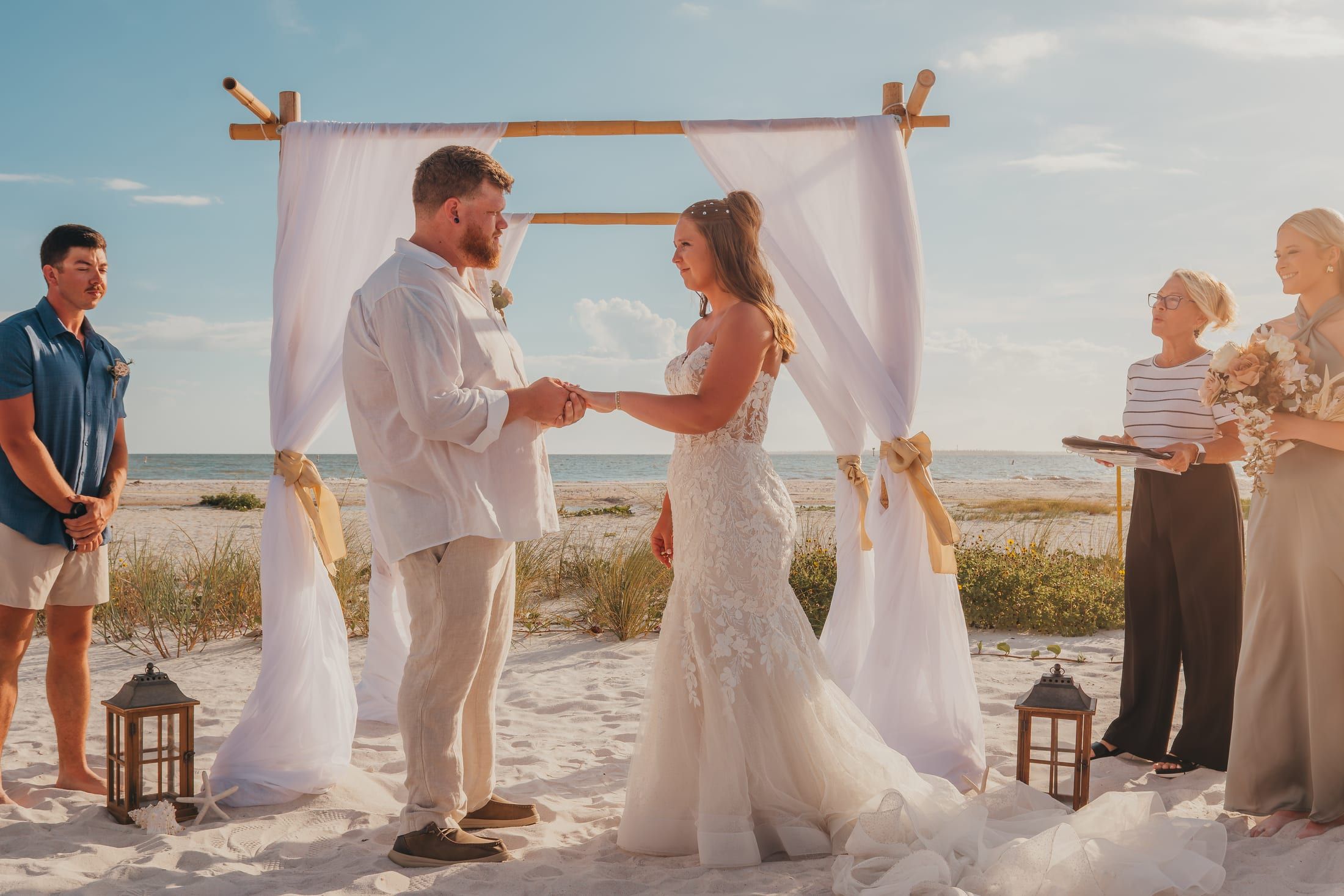 A bride and groom exchange rings at golden hour under a white draped beach arch as officiant Beth Schell leads the ceremony.