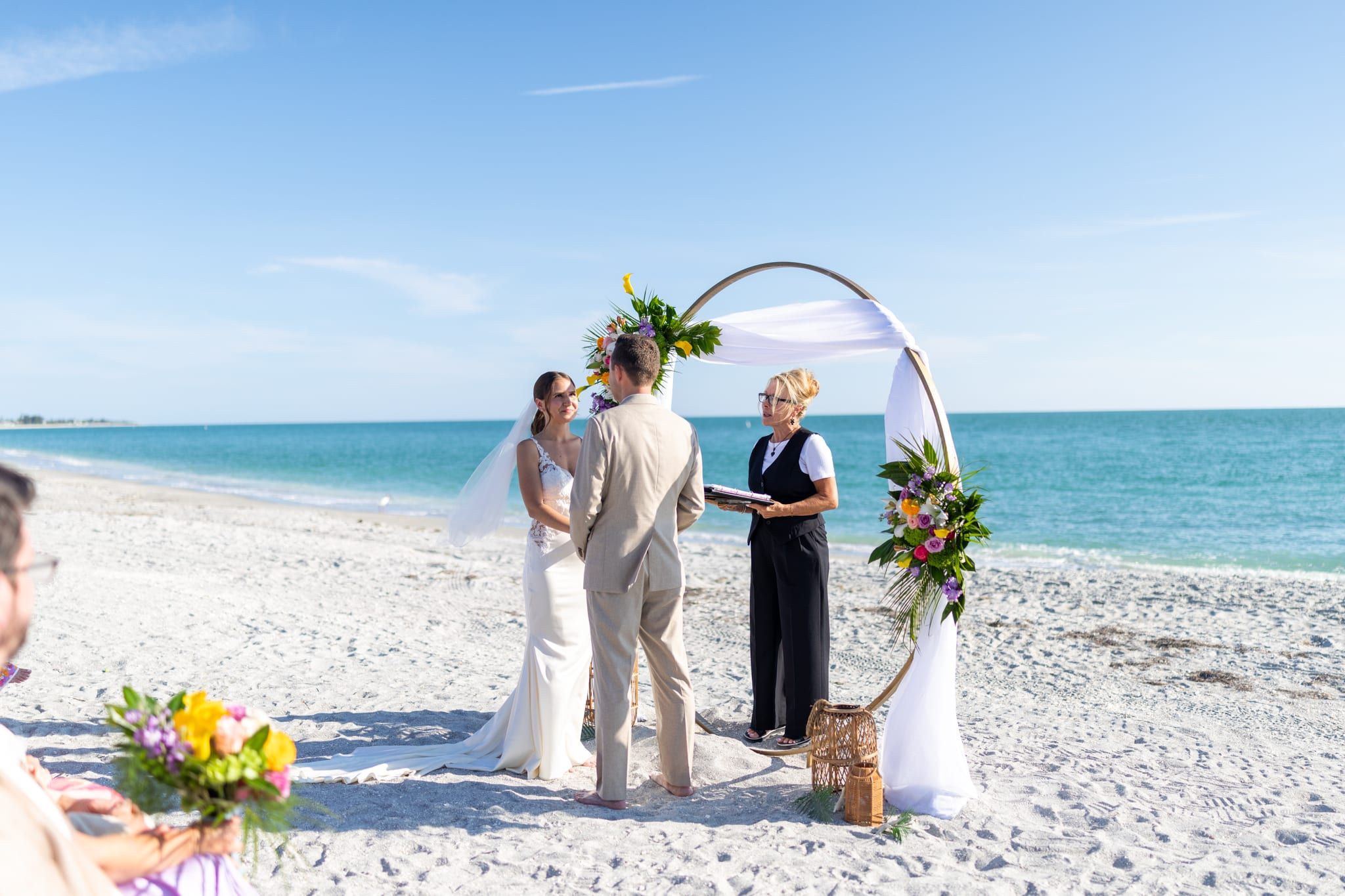 A bride and groom hold hands before a circular floral arch on a turquoise Gulf beach as officiant Beth Schell leads their ceremony.