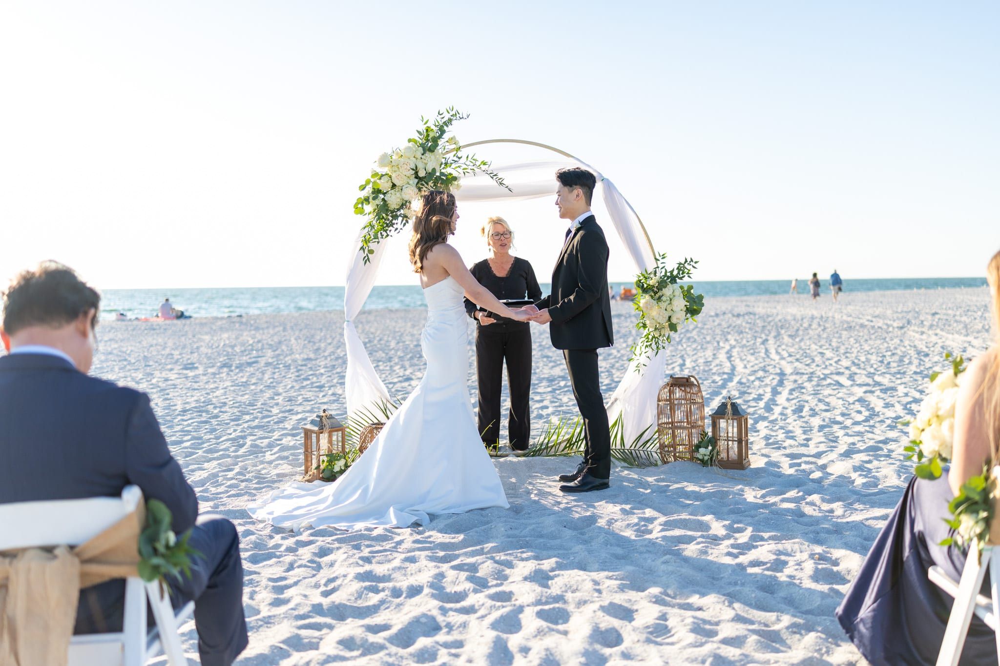 A bride and groom hold hands before a circular white floral arch on the beach as officiant Beth Schell officiates, family looking on.